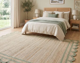 Peut inclure: Un tapis rectangulaire en fibres de jute tissées naturelles, doté d'une large bordure vert sauge et d'un bord festonné décoratif. Le tapis présente un centre beige texturé et est posé sur un parquet en bois clair.