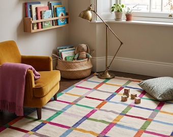 May include: A rectangular area rug featuring a cream-colored base with a grid pattern of intersecting lines in vibrant shades of blue, green, red, orange, and purple. The rug is positioned on a dark wooden floor in a room with a yellow armchair.