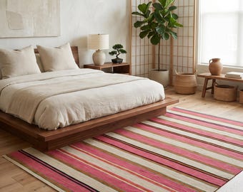 May include: A rectangular area rug featuring a repeating pattern of horizontal stripes in shades of vibrant pink, cream, dark brown, and muted green. The rug is positioned on a light-colored wooden floor in a bedroom setting, extending beneath a low wooden bed frame.