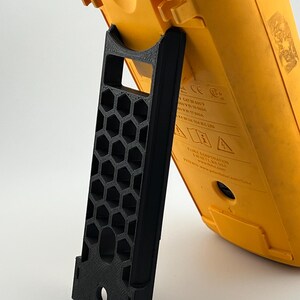 May include: A black 3D-printed stand supporting a yellow multimeter. The stand features a honeycomb pattern and a curved top to secure the device. The multimeter has a digital display and various input ports.