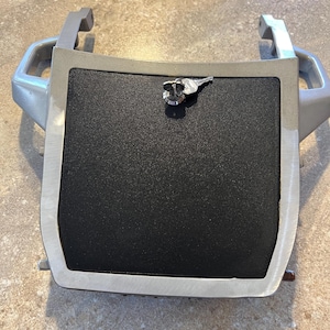 Puede incluir: Una pieza de automóvil plateada con un panel central negro texturizado. Tiene una cerradura en forma de llave y dos asas. La forma es rectangular con bordes redondeados.