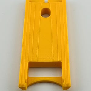 Puede incluir: Un objeto rectangular amarillo brillante con un agujero circular central y ranuras verticales. El objeto tiene una base curva y dos secciones elevadas en la parte superior. El art&iacute;culo parece estar impreso en 3D.