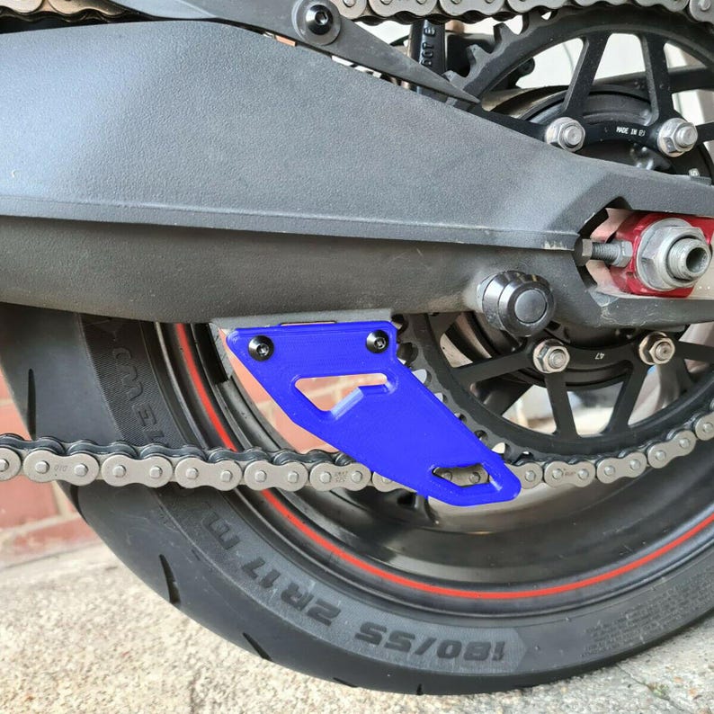Puede incluir: Un protector de cadena de motocicleta azul con forma triangular y dos orificios de montaje. El protector est&aacute; unido a una cadena de motocicleta y a un neum&aacute;tico trasero.