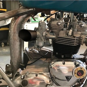 Op de afbeelding: Close-up van een motorfietsmotor met een chromen afwerking. De motor heeft een zwarte uitlaatpijp en een carburateur. De motor is gemarkeerd met "Triumph" in een zwarte driehoek.