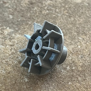 May include: A gray, 3D-printed object with a circular base and radiating blades. The object has a central hole and a cylindrical projection on one side. The design suggests a mechanical or functional component.
