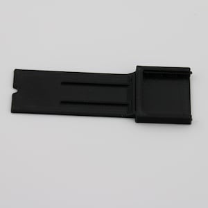 Puede incluir: Una pieza de plástico negro impresa en 3D con forma rectangular y una pequeña sección cuadrada en el lado derecho.