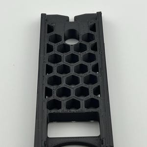 May include: A black, rectangular object with a honeycomb pattern. The object has a series of hexagonal openings and a curved bottom. The top has a notch and a circular opening. The object is made of a matte black material.