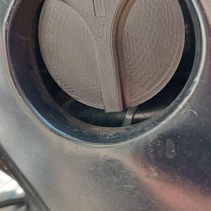 Op de afbeelding: Een close-up van een zwarte, ronde tankdop met een getextureerd oppervlak. De dop is geplaatst in een donkere, metalen behuizing. Een klein, driehoekig symbool is zichtbaar op de dop.