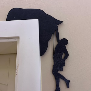 Op de afbeelding: Zwart silhouet van een persoon die op een groot object klimt, bevestigd aan een muur. De klimmer hangt aan een touw, waarbij de vorm van het object op een hoed of een wolk lijkt. De muur is lichtbeige en een wit deurkozijn is zichtbaar.