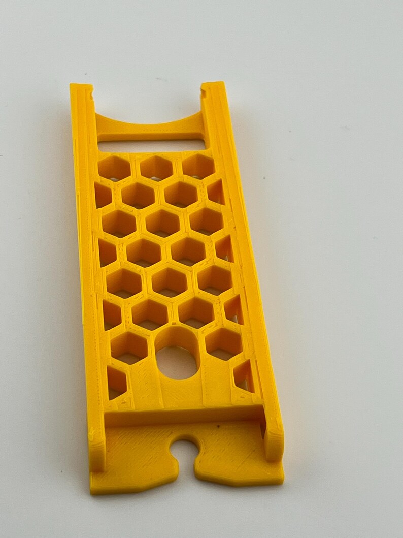 May include: A bright yellow, rectangular object with a honeycomb pattern and a curved slot at the bottom. The object has a handle at the top and is likely a tool or accessory.