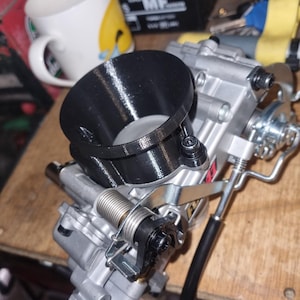 Puede incluir: Primer plano de un carburador plateado con un colector de admisión impreso en 3D negro. El carburador tiene una palanca de aceleración con resorte y una línea de combustible negra.