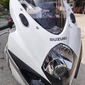 Può includere: Una moto Suzuki bianca con parabrezza e faro neri. La moto ha un design elegante con il logo Suzuki ben visibile. La carenatura anteriore è bianca con dettagli neri.