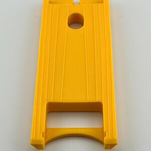 Puede incluir: Un objeto rectangular amarillo brillante con un agujero circular central y ranuras verticales. El objeto tiene una base curva y dos secciones elevadas en la parte superior. El art&iacute;culo parece estar impreso en 3D.