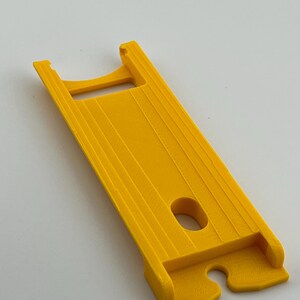 Puede incluir: Una herramienta de pl&aacute;stico amarillo brillante con un cuerpo rectangular y una abertura similar a un asa. Tiene una serie de crestas paralelas a lo largo de su longitud y un agujero circular cerca del centro. El otro extremo tiene un recorte curvo.