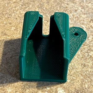 Pode incluir: Um objeto verde escuro impresso em 3D com um corpo retangular e uma aba curva. Possui uma abertura em forma de U na parte superior e um orifício na aba. A superfície tem um padrão texturizado e canelado. Parece ser um suporte.