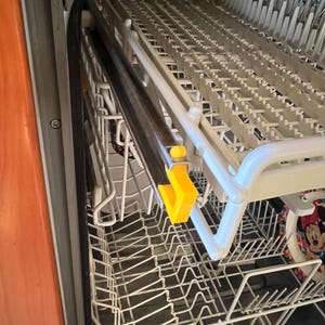 Pode incluir: Uma visão de perto da prateleira superior de uma máquina de lavar louça. A prateleira é feita de plástico branco e tem uma barra de metal que percorre a parte frontal. A prateleira está parcialmente estendida e a barra de metal é visível. A prateleira está vazia, exceto por uma pequena peça de plástico amarela que está presa à barra de metal.