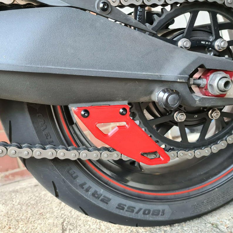 Puede incluir: Un protector de cadena de motocicleta rojo con forma triangular. El protector est&aacute; fijado a la rueda trasera de una motocicleta.