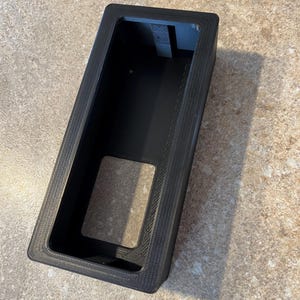 May include: A black rectangular container with a textured surface. The container has a recessed interior and a rectangular opening. The interior has a small, rectangular opening. The container is made of a dark material.