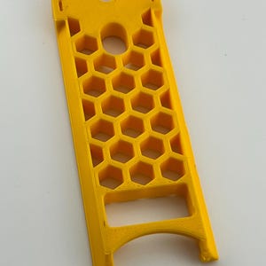 May include: A bright yellow, rectangular plastic object with a honeycomb pattern and a curved top. The object has a series of hexagonal cutouts and a central circular hole. The design includes a slot at the top and a curved base.