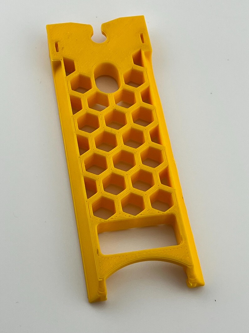 May include: A bright yellow, rectangular plastic object with a honeycomb pattern and a curved top. The object has a series of hexagonal cutouts and a central circular hole. The design includes a slot at the top and a curved base.