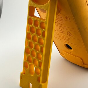 May include: A bright yellow, 3D-printed stand with a honeycomb pattern. The stand is supporting a yellow device, with text visible on the device. The stand has a slot for attachment and is designed for stability.