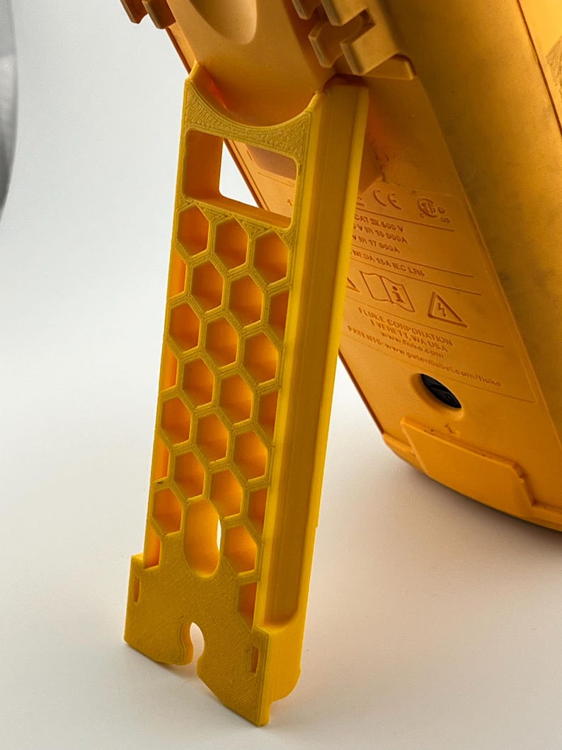 May include: A bright yellow, 3D-printed stand with a honeycomb pattern. The stand is supporting a yellow device, with text visible on the device. The stand has a slot for attachment and is designed for stability.