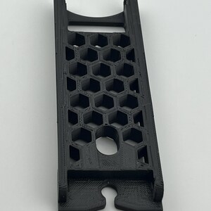 May include: A black 3D-printed phone stand with a honeycomb pattern and a circular hole. The stand has a slot at the bottom and a curved top. The stand is designed to hold a phone.