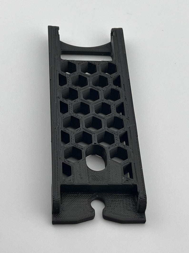 May include: A black 3D-printed phone stand with a honeycomb pattern and a circular hole. The stand has a slot at the bottom and a curved top. The stand is designed to hold a phone.