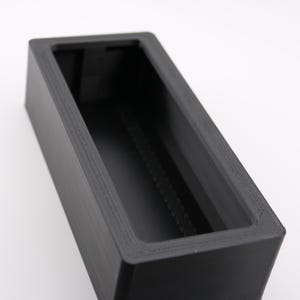 May include: A black rectangular box with a rectangular opening on the top. The box is made of a smooth, textured material.