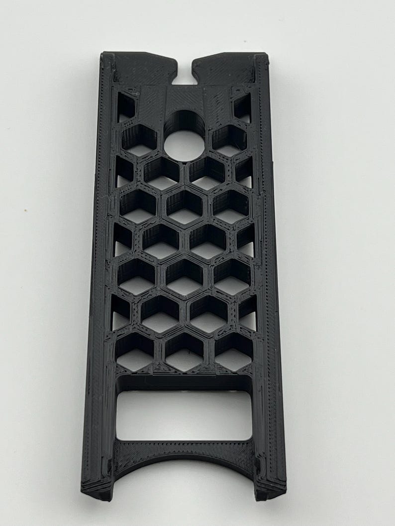 May include: A black, rectangular 3D-printed object with a honeycomb pattern. The object has a curved bottom and a notched top with a circular hole. The design features a series of hexagonal cells, creating an open, geometric structure.