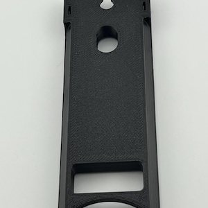 Puede incluir: Un objeto negro rectangular impreso en 3D con una ranura en la parte superior y un agujero circular en el centro. El objeto tiene una abertura rectangular en la parte inferior y una superficie texturizada.
