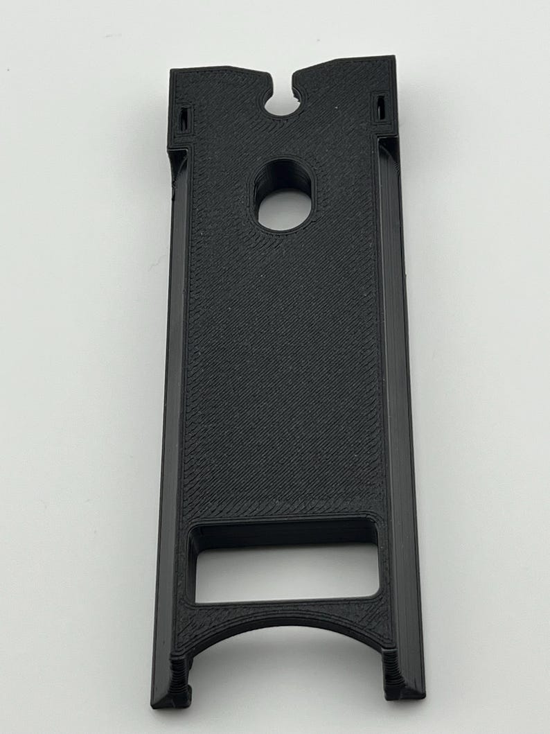 Puede incluir: Un objeto negro rectangular impreso en 3D con una ranura en la parte superior y un agujero circular en el centro. El objeto tiene una abertura rectangular en la parte inferior y una superficie texturizada.