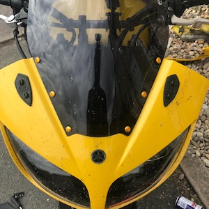 Może przedstawiać: Zbliżenie na żółty motocykl z czarną szybą i logo Yamaha. Motocykl ma czarną lampę przednią i kilka złotych śrub.