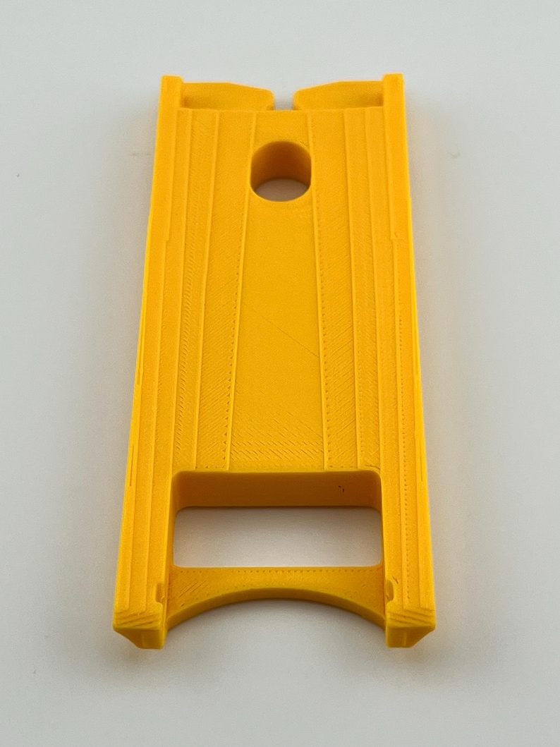 Puede incluir: Un objeto rectangular amarillo brillante con un agujero circular central y ranuras verticales. El objeto tiene una base curva y dos secciones elevadas en la parte superior. El art&iacute;culo parece estar impreso en 3D.