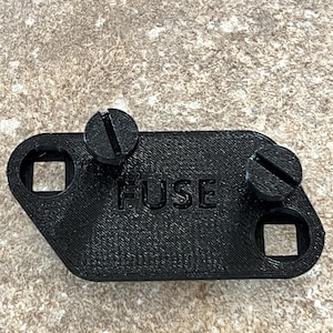 Könnte beinhalten: Ein schwarzer Sicherungshalter aus Kunststoff mit dem Wort "FUSE" auf der Oberfläche. Zwei schwarze Schrauben sind angebracht. Der Halter hat an beiden Enden quadratische Löcher. Der Hintergrund ist ein gesprenkeltes Braun und Beige.