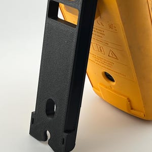 Puede incluir: Un soporte negro impreso en 3D que sostiene un dispositivo electr&oacute;nico amarillo. El soporte tiene forma rectangular con un recorte y un asa. El dispositivo tiene texto y s&iacute;mbolos.