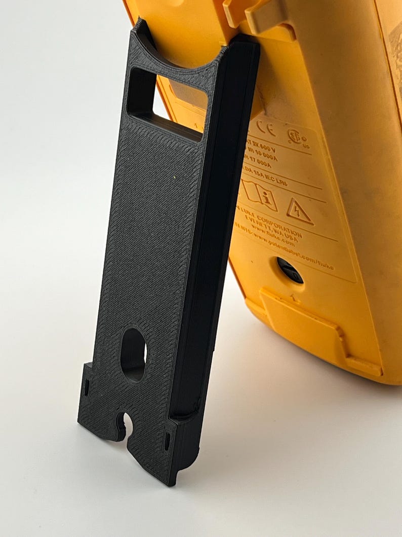 Puede incluir: Un soporte negro impreso en 3D que sostiene un dispositivo electr&oacute;nico amarillo. El soporte tiene forma rectangular con un recorte y un asa. El dispositivo tiene texto y s&iacute;mbolos.