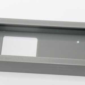 Puede incluir: Una caja rectangular de plástico gris con una abertura rectangular en la parte delantera. La caja tiene una superficie texturizada.