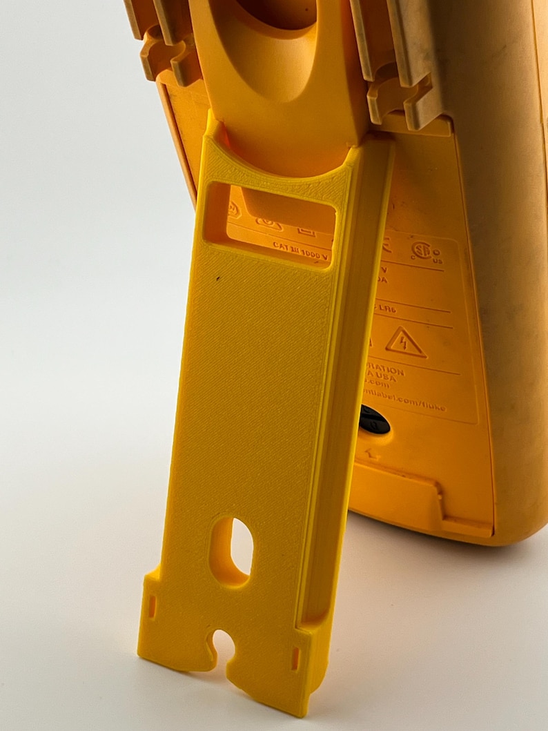 Puede incluir: Primer plano de un dispositivo de prueba el&eacute;ctrica amarillo con un soporte incorporado. El soporte es de color amarillo brillante y tiene forma rectangular con un recorte curvo. El dispositivo tiene el texto "CAT III 1000 V".