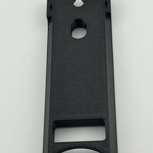 Puede incluir: Un objeto negro rectangular impreso en 3D con una ranura en la parte superior y un agujero circular en el centro. El objeto tiene una abertura rectangular en la parte inferior y una superficie texturizada.