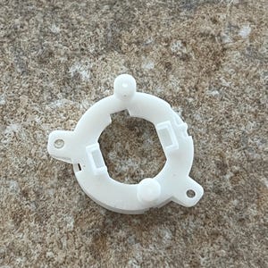 Könnte beinhalten: Eine weiße Kunststoff-Kreiskomponente mit einer zentralen Öffnung und mehreren hervorstehenden Merkmalen. Die Komponente hat eine ringartige Form mit zwei abgerundeten Vorsprüngen und zwei rechteckigen Aussparungen. Sie hat auch zwei kleine Löcher.