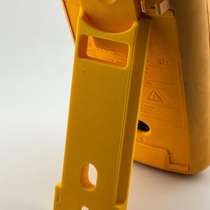 Puede incluir: Primer plano de un dispositivo de prueba el&eacute;ctrica amarillo con un soporte incorporado. El soporte est&aacute; extendido, mostrando una abertura rectangular y un recorte curvo. El dispositivo tiene el texto "CAT III 1000 V".