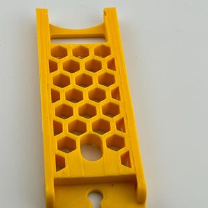 May include: A bright yellow, rectangular object with a honeycomb pattern and a curved slot at the bottom. The object has a handle at the top and is likely a tool or accessory.