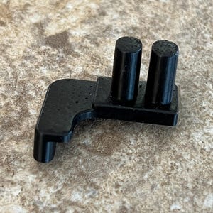 May include: A small, black plastic component featuring two cylindrical posts and a right-angled extension. The object appears 3D-printed and has a textured surface. The component is likely part of a larger mechanism or device.
