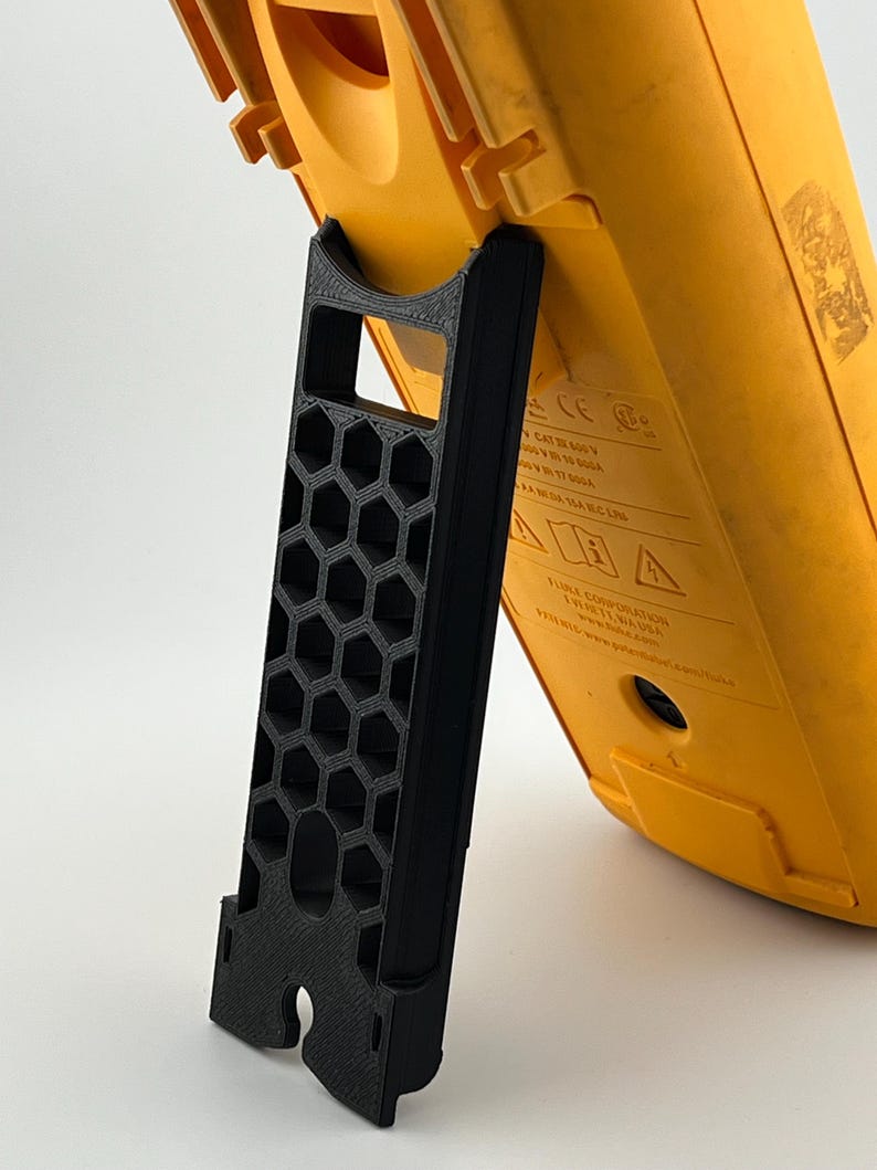 May include: A black 3D-printed stand supporting a yellow multimeter. The stand features a honeycomb pattern and a curved top to secure the device. The multimeter has a digital display and various input ports.