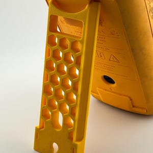 May include: Close-up of a yellow digital multimeter with a honeycomb-patterned stand. The stand is angled, providing support. The multimeter has various labels and symbols, including voltage and amperage ratings.