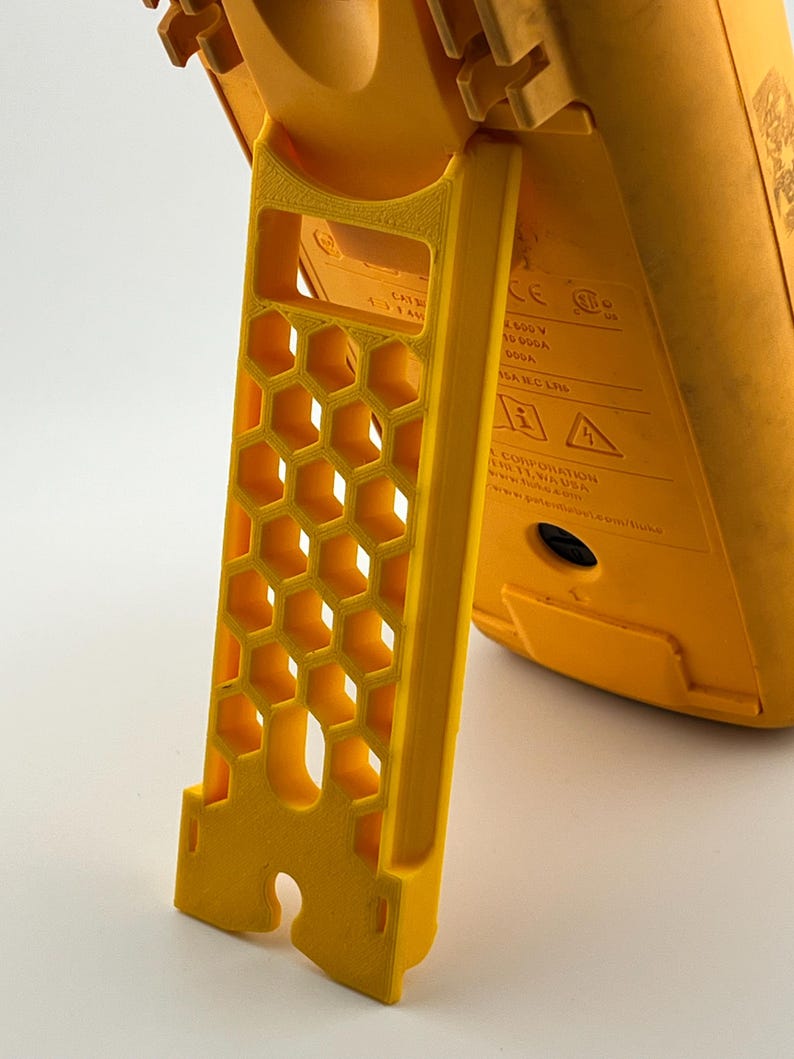 May include: Close-up of a yellow digital multimeter with a honeycomb-patterned stand. The stand is angled, providing support. The multimeter has various labels and symbols, including voltage and amperage ratings.