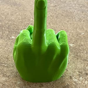 May include: A vivid green, 3D-printed hand sculpture with the middle finger raised. The sculpture is a single colour and shows layer lines from the printing. The hand is positioned on a neutral-toned surface.