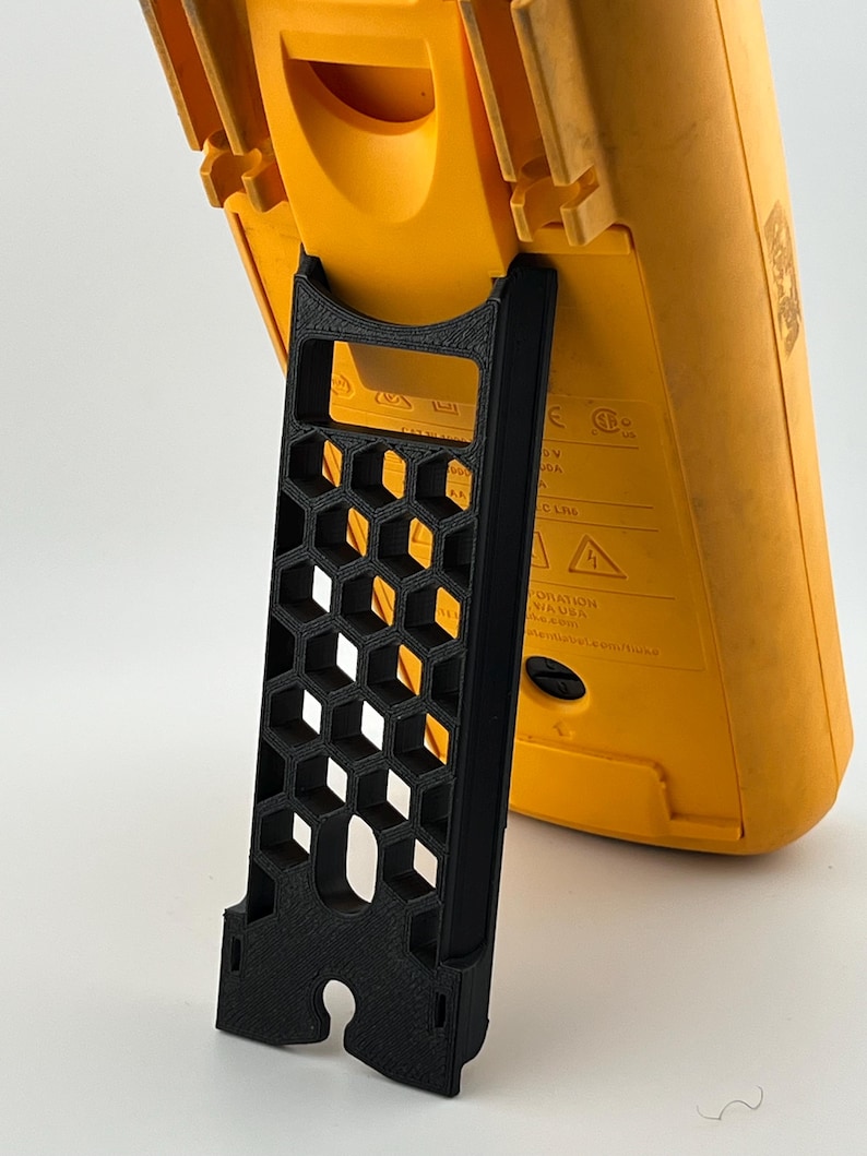 May include: A black 3D-printed stand with a honeycomb design, supporting a yellow multimeter. The stand has a rectangular shape with a curved base and a series of hexagonal cutouts. The multimeter is yellow with black text and markings.