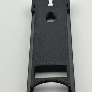 Puede incluir: Un objeto negro rectangular impreso en 3D con una superficie texturizada. Presenta un agujero circular cerca de la parte superior y una abertura curva en la parte inferior. El dise&ntilde;o incluye una ranura en la parte superior y una abertura rectangular.